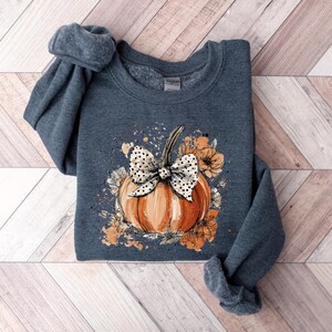 May include: A dark gray sweatshirt featuring a watercolor design of an orange pumpkin with a white polka dot bow and floral accents. The pumpkin is the focal point, surrounded by orange and cream-colored flowers. The sleeves are rolled up.