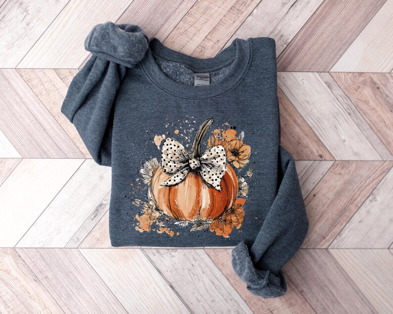 May include: A dark gray sweatshirt featuring a watercolor design of an orange pumpkin with a white polka dot bow and floral accents. The pumpkin is the focal point, surrounded by orange and cream-colored flowers. The sleeves are rolled up.