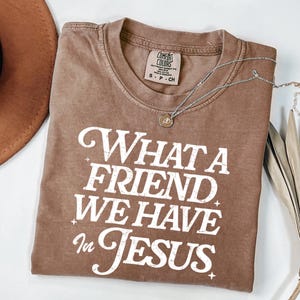K&ouml;nnte beinhalten: Ein braunes T-Shirt mit einem wei&szlig;en, abgenutzten Aufdruck, der "What a Friend We Have in Jesus" lautet.