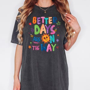 May include: Dark gray t-shirt with the text "BETTER DAYS ARE ON THE WAY" in colorful letters. The design includes a sun, flowers, bees, and hearts. The shirt has short sleeves and a relaxed fit.