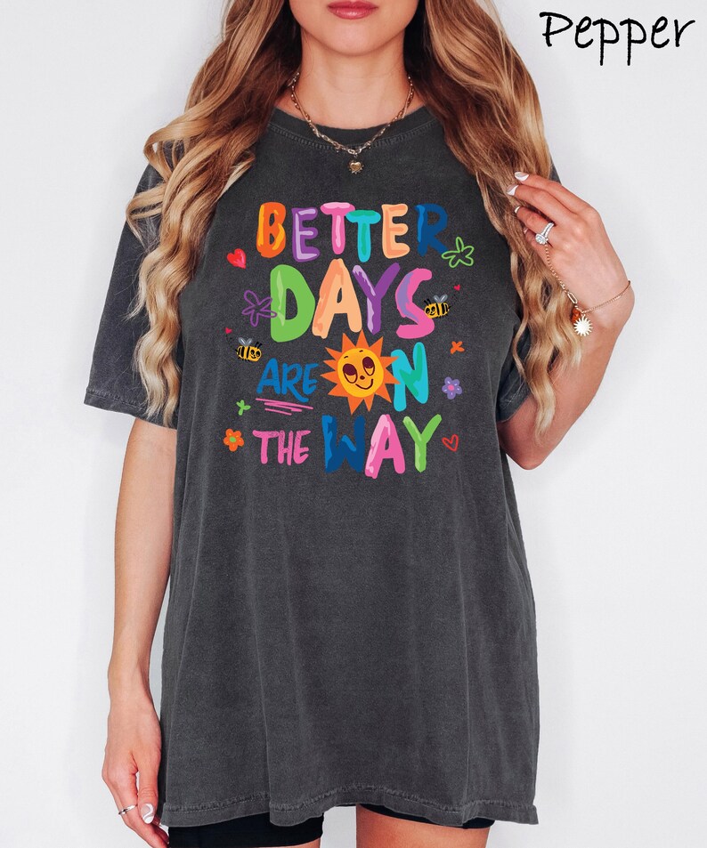 May include: Dark gray t-shirt with the text "BETTER DAYS ARE ON THE WAY" in colorful letters. The design includes a sun, flowers, bees, and hearts. The shirt has short sleeves and a relaxed fit.