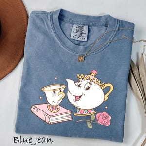 May include: Blue jean colored t-shirt featuring a cartoon illustration of Mrs. Potts, Chip, a book, and a rose. The shirt has a crew neck and short sleeves. A gold necklace is draped across the shirt. The text "Blue Jean" is at the bottom.