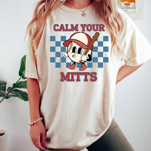 May include: A white t-shirt with a baseball graphic and the text "Calm Your Mitts". The graphic features a cartoon baseball with a red hat, a blue checkered background, and a baseball bat.