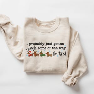 May include: Cream-colored sweatshirt with the text "probably just gonna jingle some of the way I'm tired." The graphic features four dachshunds dressed in holiday attire. The sleeves are rolled up.