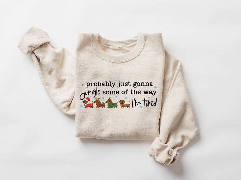 May include: Cream-colored sweatshirt with the text "probably just gonna jingle some of the way I'm tired." The graphic features four dachshunds dressed in holiday attire. The sleeves are rolled up.