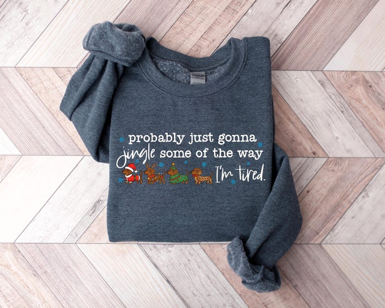 May include: A dark gray sweatshirt with the text "probably just gonna jingle some of the way I'm tired." The graphic includes cartoon dachshunds dressed as Santa, reindeer, and a Christmas tree. The sweatshirt is folded on a light wood surface.