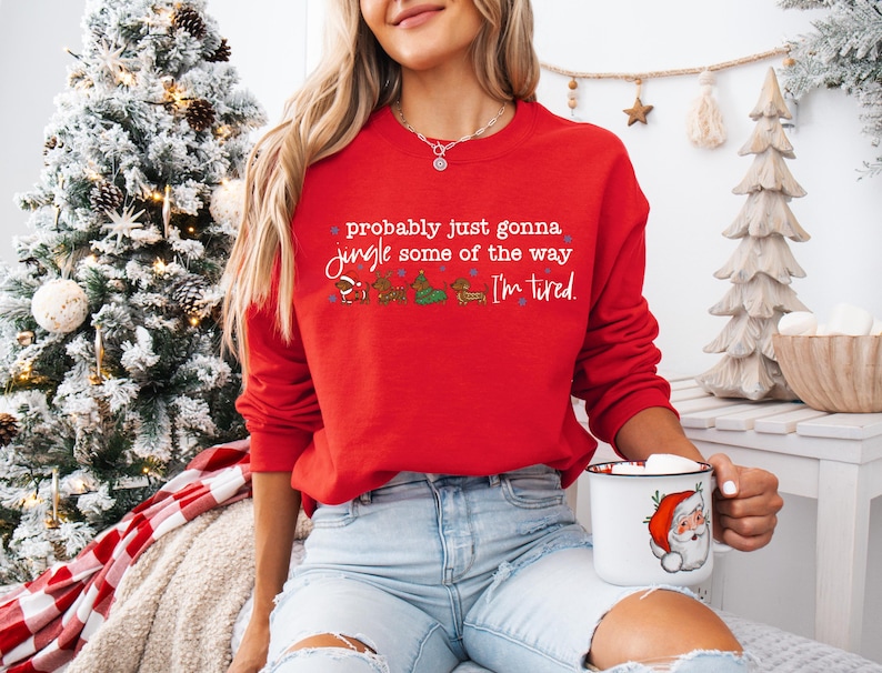 May include: A red sweatshirt with the text "probably just gonna jingle some of the way, I'm tired." The sweatshirt has a festive design with cartoon characters. A white mug with a Santa Claus graphic is held in the hand.