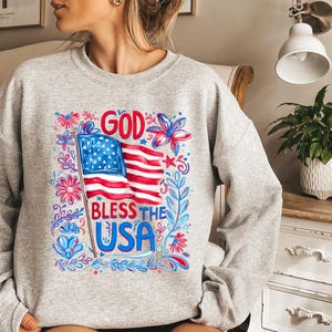 May include: A light grey crewneck sweatshirt featuring a graphic print of the American flag with the text GOD BLESS THE USA in red and blue lettering, surrounded by stylized red and blue floral and leaf illustrations on the front center.