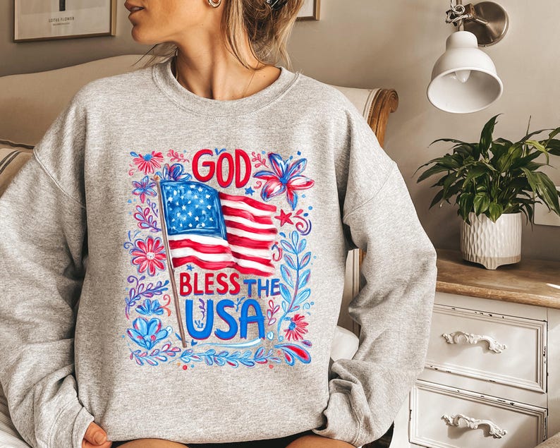 May include: A light grey crewneck sweatshirt featuring a graphic print of the American flag with the text GOD BLESS THE USA in red and blue lettering, surrounded by stylized red and blue floral and leaf illustrations on the front center.
