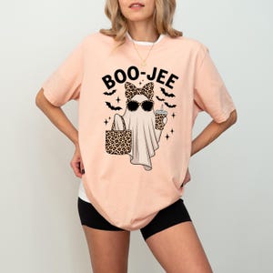 May include: Peach-coloured t-shirt featuring a ghost graphic wearing sunglasses and a leopard print bow, carrying a leopard print tote bag and a drink. The text "BOO-JEE" is above the ghost, with bats and stars around it.