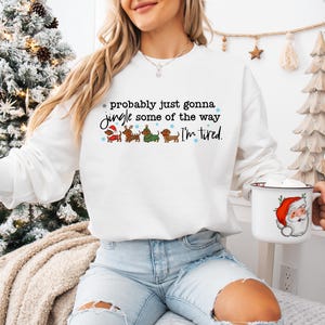 May include: White sweatshirt with the text "probably just gonna jingle some of the way I'm tired." The sweatshirt features dachshunds in Christmas outfits. A white mug with a Santa Claus graphic is held in the hand.