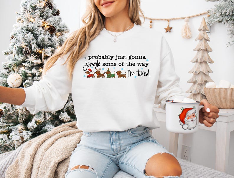 May include: White sweatshirt with the text "probably just gonna jingle some of the way I'm tired." The sweatshirt features dachshunds in Christmas outfits. A white mug with a Santa Claus graphic is held in the hand.