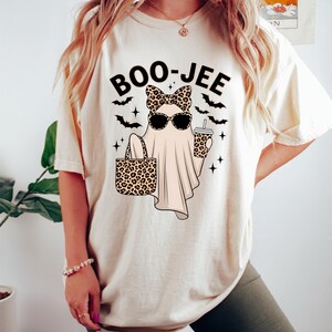 May include: Cream-coloured t-shirt featuring a ghost graphic wearing sunglasses and a leopard print bow, holding a leopard print tote bag and a drink. The text "BOO-JEE" is above the ghost, with bats and stars.