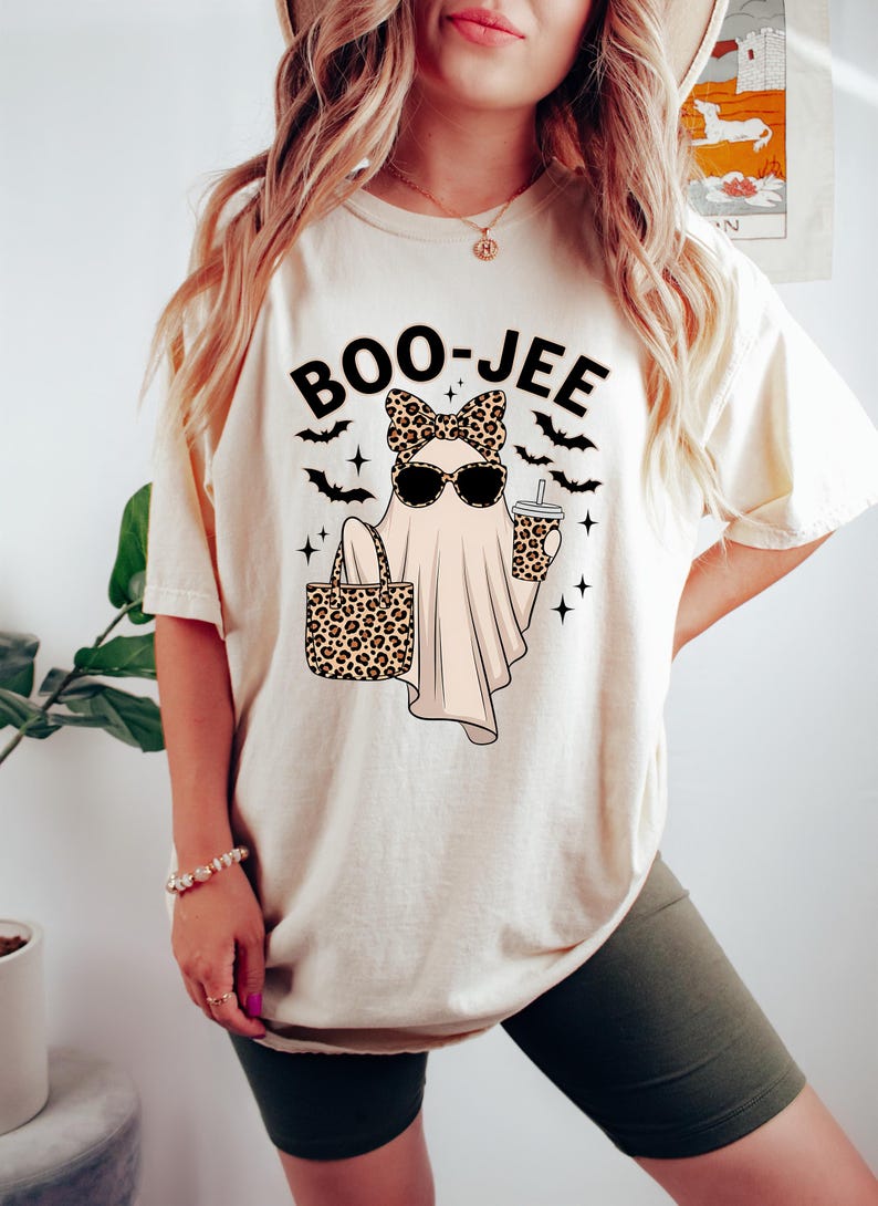 May include: Cream-coloured t-shirt featuring a ghost graphic wearing sunglasses and a leopard print bow, holding a leopard print tote bag and a drink. The text "BOO-JEE" is above the ghost, with bats and stars.