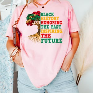 Pu&ograve; includere: T-shirt rosa con la scritta "BLACK HISTORY HONORING THE PAST INSPIRING THE FUTURE" in nero, rosso, giallo e verde. La maglietta presenta una grafica ad albero con foglie colorate e radici.
