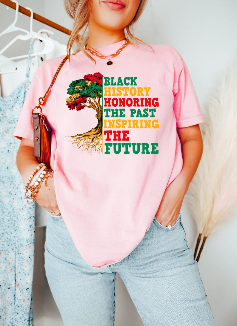 Pu&ograve; includere: T-shirt rosa con la scritta "BLACK HISTORY HONORING THE PAST INSPIRING THE FUTURE" in nero, rosso, giallo e verde. La maglietta presenta una grafica ad albero con foglie colorate e radici.