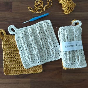 Puede incluir: Dos paños de cocina de ganchillo, uno de color beige con un patrón de punto de cable y el texto "HANDCRAFTED Kitchen Cloth" y el otro de color amarillo mostaza con un patrón de punto simple.