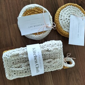 Puede incluir: Conjunto de tres posavasos de crochet con ribete amarillo mostaza y un paño de cocina de crochet blanco con el texto "HANDCRAFTED Kitchen Cloth".