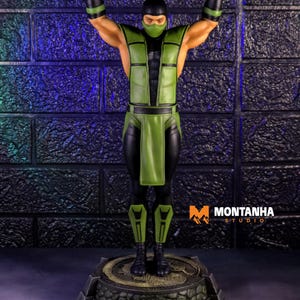 Puede incluir: Figura detallada de un personaje con un atuendo verde y negro, con los brazos levantados. La figura tiene una máscara y está de pie sobre una base circular con un diseño de dragón. El fondo es una pared de ladrillos texturizada.