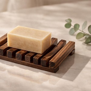 May include: A rectangular bar of light brown soap rests on a dark brown wooden soap dish. The dish has a slatted design. A sprig of green eucalyptus is in the background. The soap dish is approximately 5 inches long.
