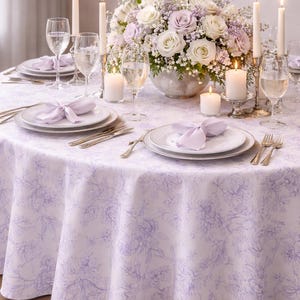 May include: A beautifully set dining table with a white and lavender floral patterned tablecloth. The table is adorned with plates, silverware, glasses, and lit candles. A floral centerpiece of white and lavender roses adds elegance.