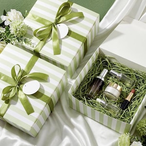 May include: Two square gift boxes with light green and white stripes, tied with olive green satin ribbons and blank white circular tags. One box is open, revealing cosmetic bottles, a cream jar, and lipstick nestled in green paper shred.