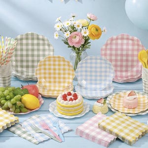 May include: A festive picnic scene with gingham plates, cups, and napkins in pastel colors. The table is set with fruit, cake, and colorful cutlery. Balloons and flowers add to the celebratory atmosphere, perfect for a party or outdoor gathering.