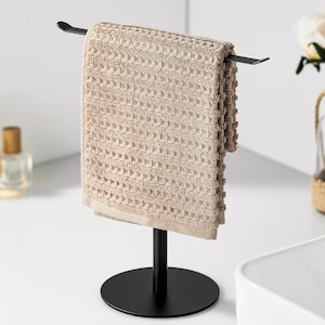 May include: A beige waffle-weave towel hangs on a black metal towel stand. The stand has a circular base and a vertical post that supports the towel bar. The towel is folded and has a textured surface. The stand is a modern bathroom accessory.