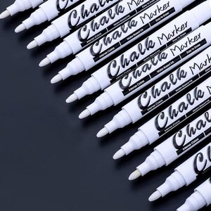 May include: A collection of white chalk markers with black text on a dark background. The markers are labeled "Chalk Marker" and "Intense Colors". The markers have a conical tip and are arranged diagonally.