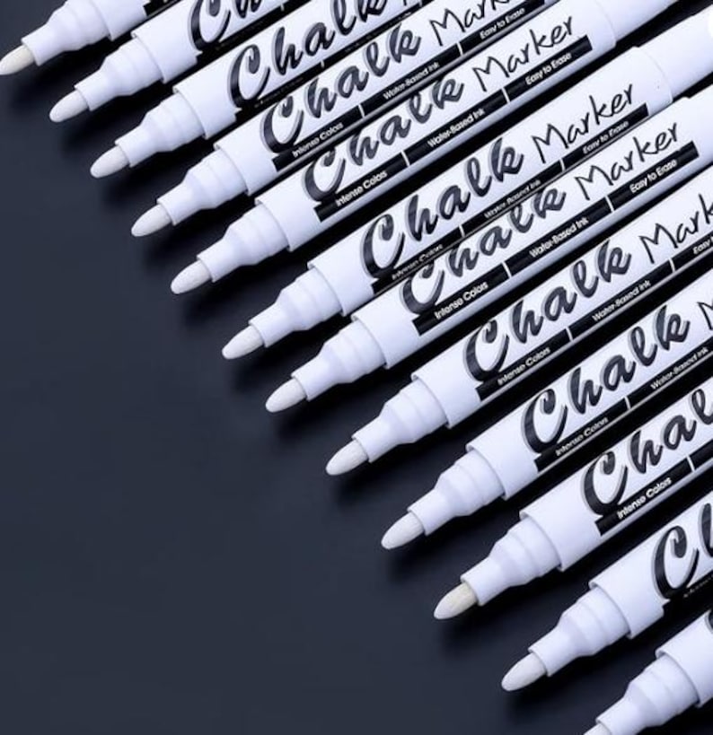 May include: A collection of white chalk markers with black text on a dark background. The markers are labeled "Chalk Marker" and "Intense Colors". The markers have a conical tip and are arranged diagonally.