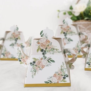 May include: White decorative teapot-shaped boxes with floral designs in soft pink, peach, and green. The boxes have gold trim and are arranged on a white surface. The teapots have a crown-like top and a handle.