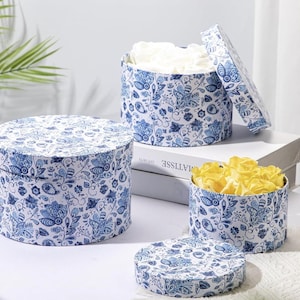 May include: Three round gift boxes with lids, decorated with a blue and white floral pattern. One box is open, revealing white flowers, while another contains yellow flowers. The boxes are arranged on a white surface.
