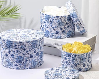 May include: Three round gift boxes with lids, decorated with a blue and white floral pattern. One box is open, revealing white flowers, while another contains yellow flowers. The boxes are arranged on a white surface.