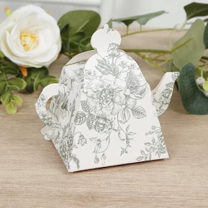 May include: A white teapot-shaped gift box with a floral pattern in gray. The box has a handle, spout, and a crown-like detail on the lid. The background includes white flowers and green leaves.