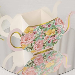 May include: A decorative paper teapot with a floral pattern of pink roses and yellow flowers on a teal background. The teapot has a pink handle and spout with gold trim. The design is reflected on a mirrored surface.