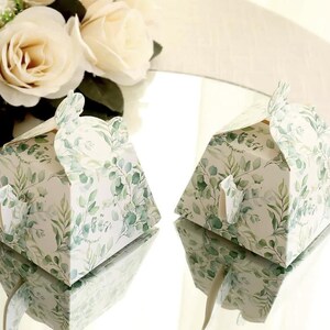 May include: Two white, hexagonal gift boxes with a green and white botanical print. The boxes have a flap closure and a decorative leaf-shaped accent. A bouquet of white roses is in the background.