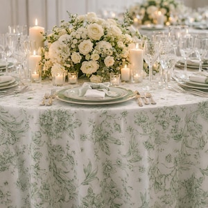 May include: Elegant table setting with a white and green floral tablecloth. The table is set with plates, silverware, and wine glasses. White candles and floral arrangements add to the ambiance. A perfect setting for a special occasion.