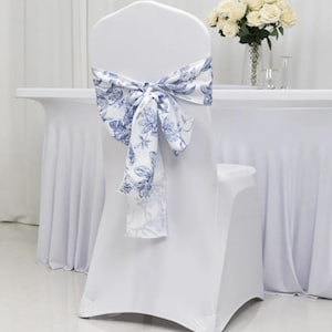 May include: A white chair cover with a blue and white floral patterned bow. The bow is tied at the back of the chair. A vase of white roses sits on a white table in the background.