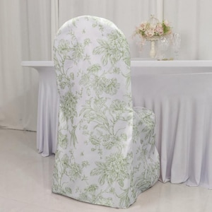 May include: A white chair cover with a green floral pattern. The cover is draped over a chair, with a table and floral arrangement in the background. The floral design features leaves and flowers.