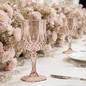 May include: Pink-tinted stemmed glassware with diamond-patterned bowls and slender stems. The glasses are set on a white tablecloth, with a blurred background of pink flowers. A knife and fork are visible.