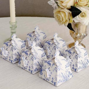 May include: Six white gift boxes with a blue floral pattern are arranged on a white surface. The boxes have a unique shape with a decorative top closure. A bouquet of cream-colored roses and a candle holder are in the background.