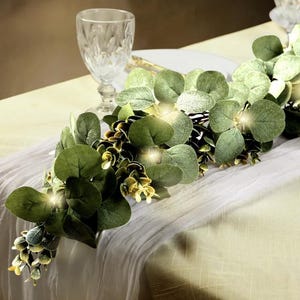 May include: A decorative eucalyptus garland with large, round green leaves and small yellow accents. The garland is draped across a white tablecloth, with a crystal wine glass in the background. Small lights are interspersed throughout the garland.