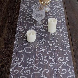 May include: A decorative table runner with a gray wood grain pattern and a white, swirling floral design. Two white pillar candles and a clear glass are on the runner. The runner is approximately 12 inches wide.