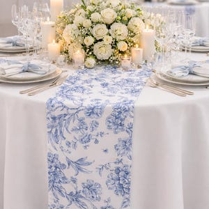 May include: Elegant table setting with a white tablecloth, blue floral runner, and white roses centerpiece. Includes candles, glassware, and plates with blue ribbon accents, creating a sophisticated ambiance for a special event.