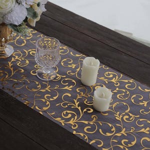 May include: A gray table runner with a gold floral pattern, a clear glass, and two white candles. The runner is placed on a dark wooden table. A vase of white and yellow flowers is visible in the background.