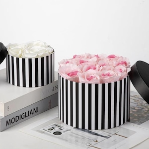 May include: Two round flower boxes with black and white vertical stripes. One box is filled with white roses, the other with pink roses. The boxes have black lids and sit on a white surface.