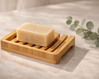 May include: A rectangular bar of soap rests on a bamboo soap dish. The soap is light beige, and the dish has a slatted design. A sprig of eucalyptus adds a natural touch. The soap dish is approximately 5.5 inches long.