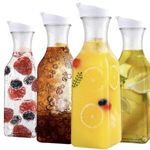 May include: Four clear glass carafes with white lids, each filled with a different beverage. One contains berries, another a brown liquid with bubbles, a third has orange slices, and the last has lemon slices.