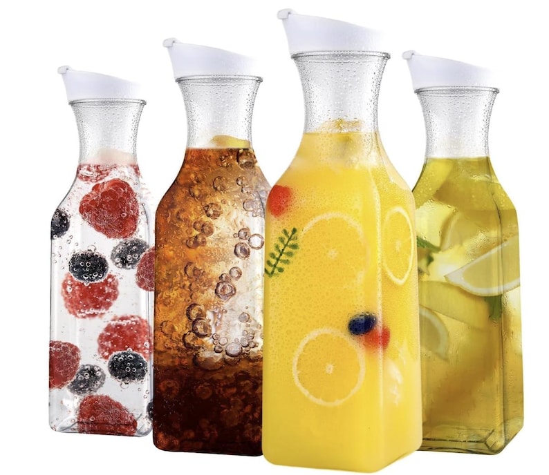 May include: Four clear glass carafes with white lids, each filled with a different beverage. One contains berries, another a brown liquid with bubbles, a third has orange slices, and the last has lemon slices.