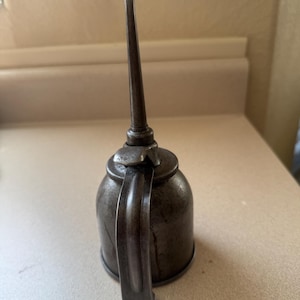 May include: A vintage metal oil can with a dark, weathered patina. The object features a rounded base, a curved handle attached to the side, and a long, slender, tapered spout extending vertically from the top, accompanied by a small thumb lever.